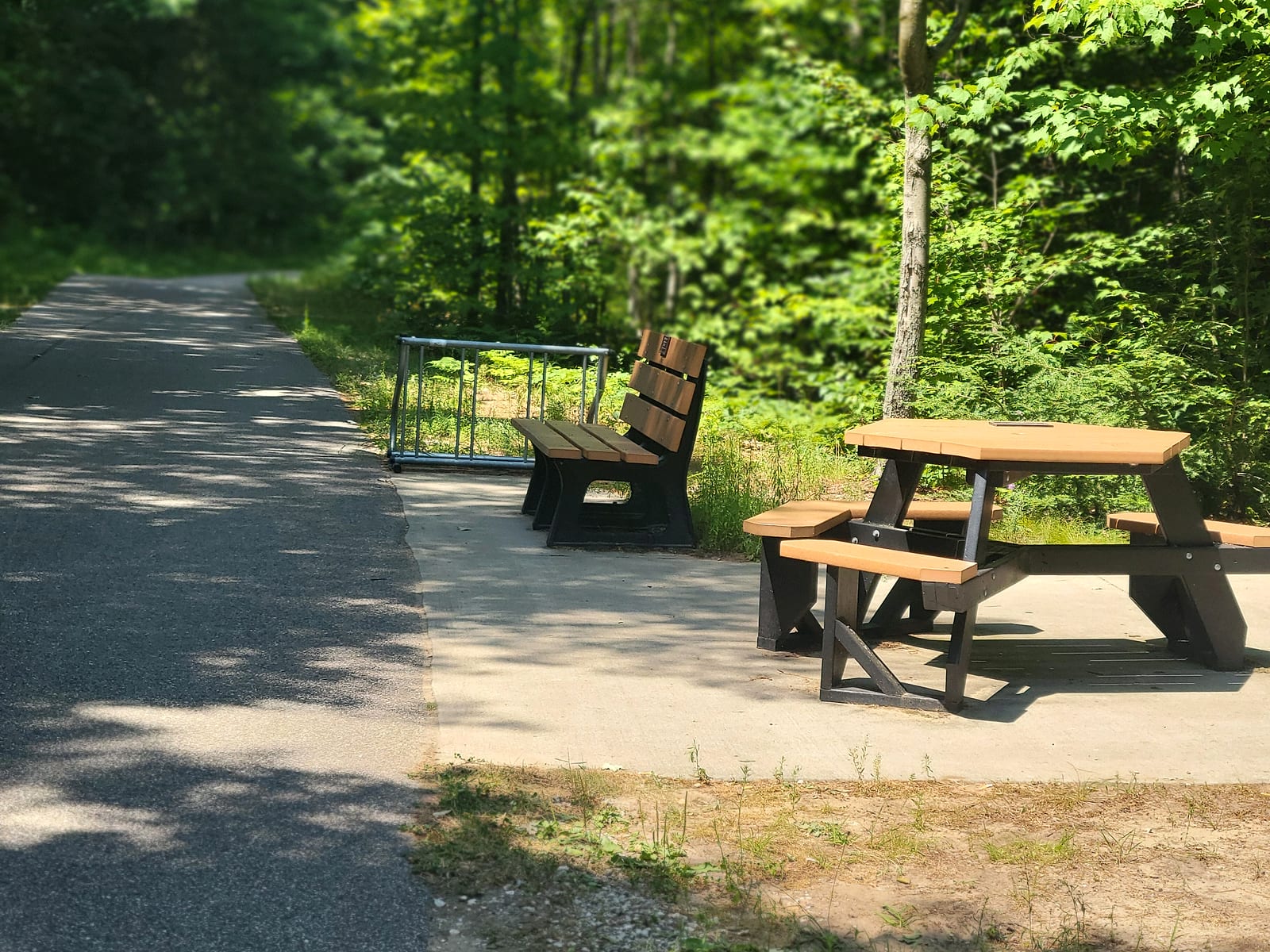 Bench, picnic table, and bike rack on a concrete rest pad beside the paved Berry Junction Trail in the woods north of Muskegon