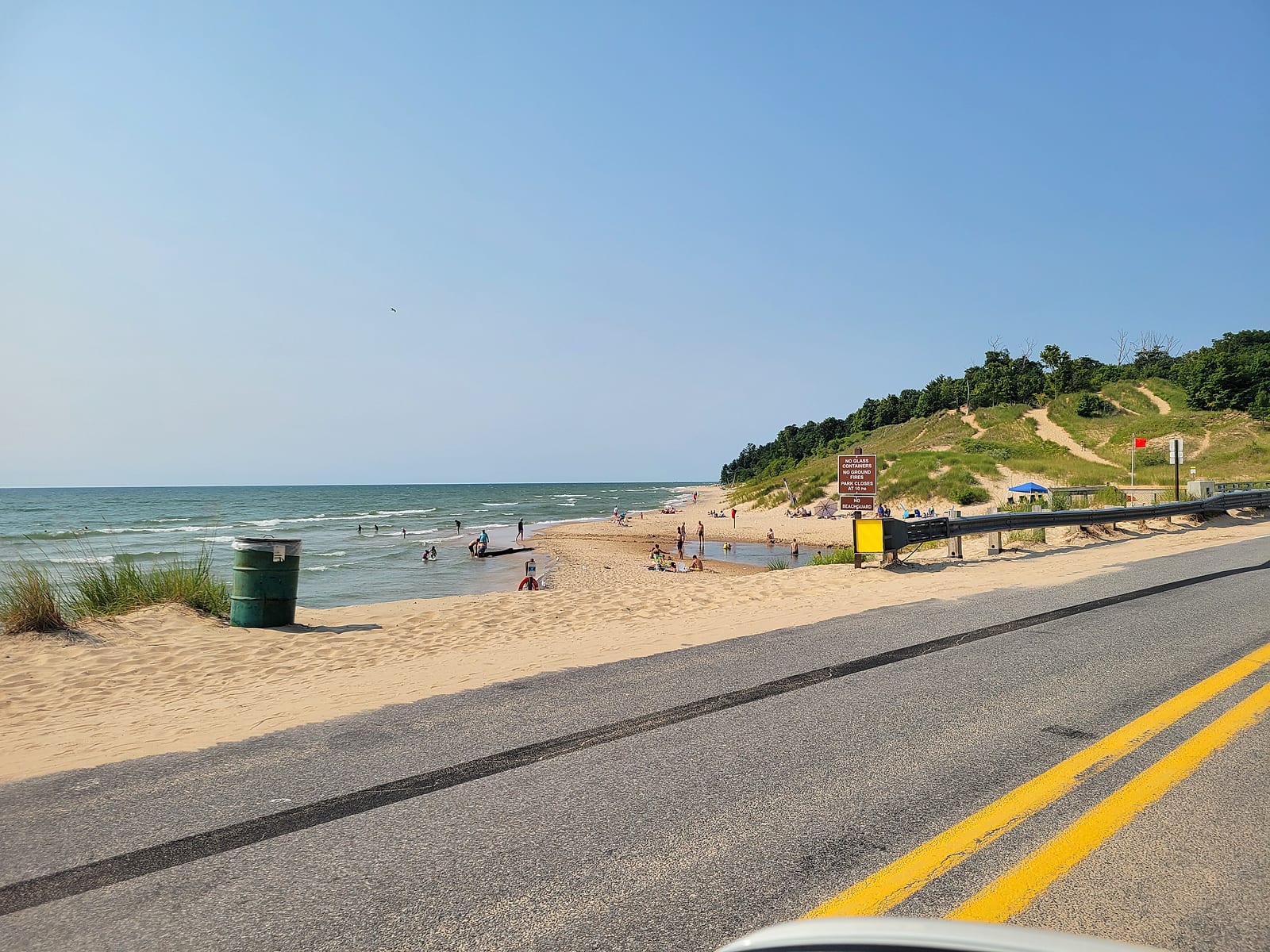 The Duck Lake outlet stream braiding across the sandy Lake Michigan beach at Duck Lake State Park, with beachgoers, dunes, and the posted park rules sign