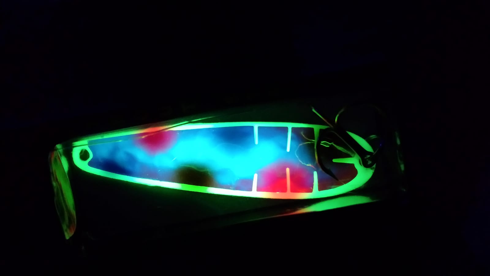 A glow salmon trolling spoon photographed in the dark, glowing green around a blue and pink pattern, treble hook at the tail