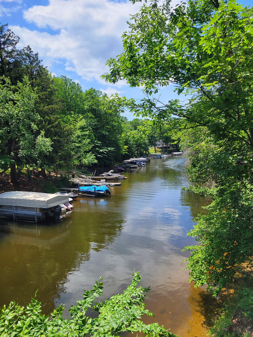 The narrow, wooded Mona Lake channel with pontoon boats docked along the bank