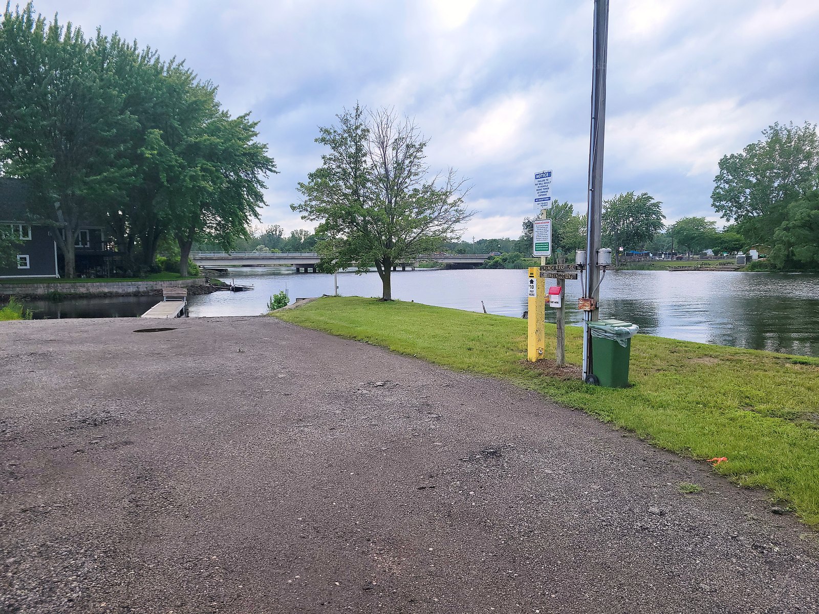A private boat launch ramp on N. Causeway Street descending into the North Branch of the Muskegon River, with a yellow ten-dollar pay post and a launch-use instructions sign in the foreground and the low causeway bridge crossing the river in the background.
