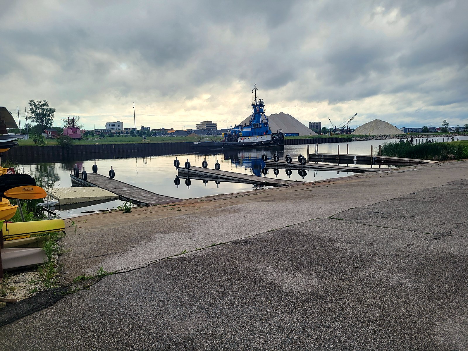 Fisherman's Landing boat launch at the east, river end of Muskegon Lake, with floating courtesy docks, a wide concrete ramp, and a moored tug across the channel beside industrial aggregate piles.