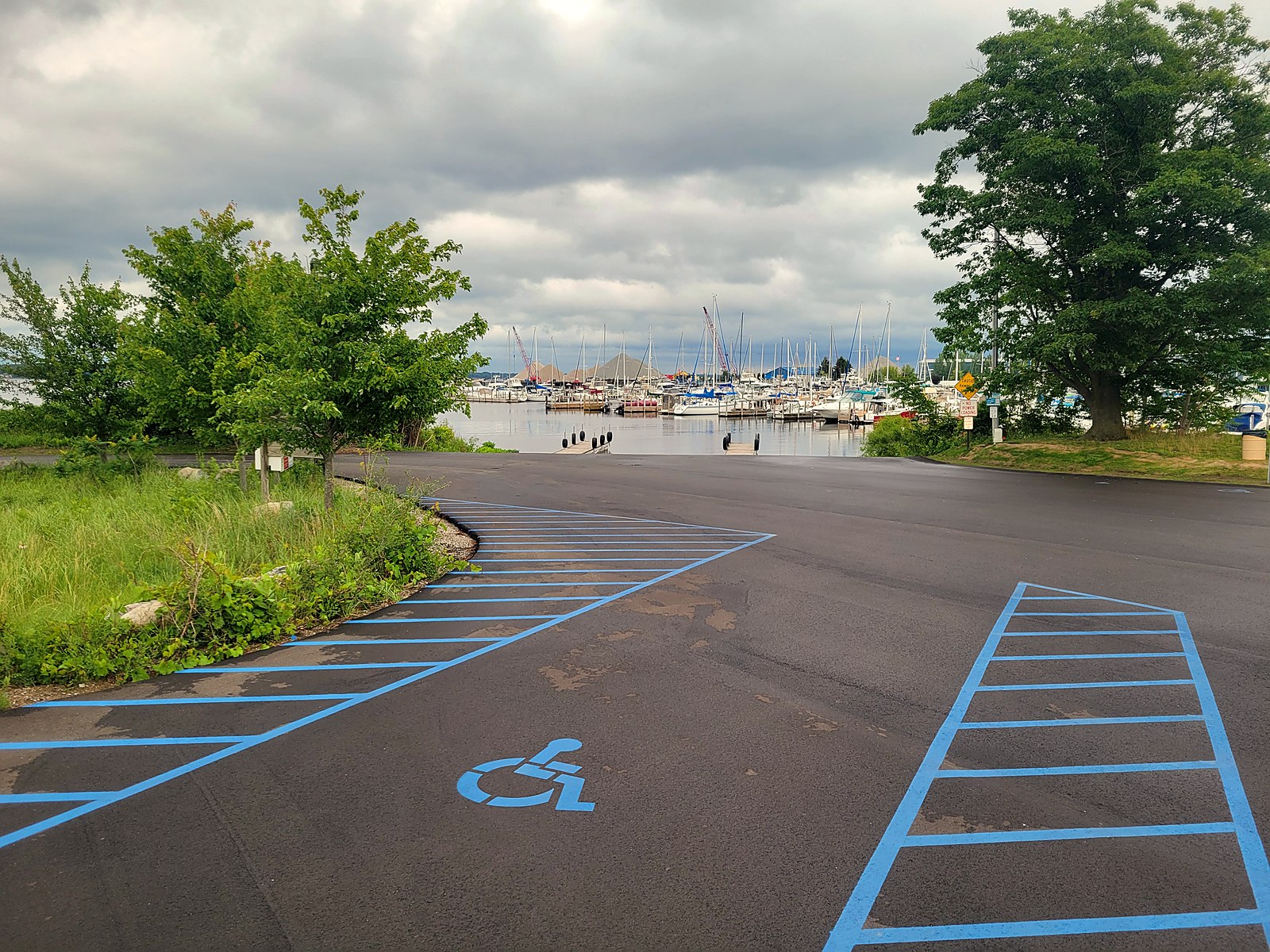 The Grand Trunk launch ramp and its large paved parking lot on the south shore of Muskegon Lake, with fresh blue accessible-parking striping in the foreground and a marina full of moored sailboats across the channel under an overcast sky.