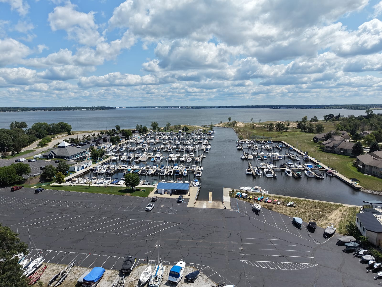 July 2026 aerial drone view of Harbour Towne Marina in Muskegon: the public boat launch ramp and courtesy dock between two fields of slips, trailer parking in the foreground, and the basin opening onto Muskegon Lake.