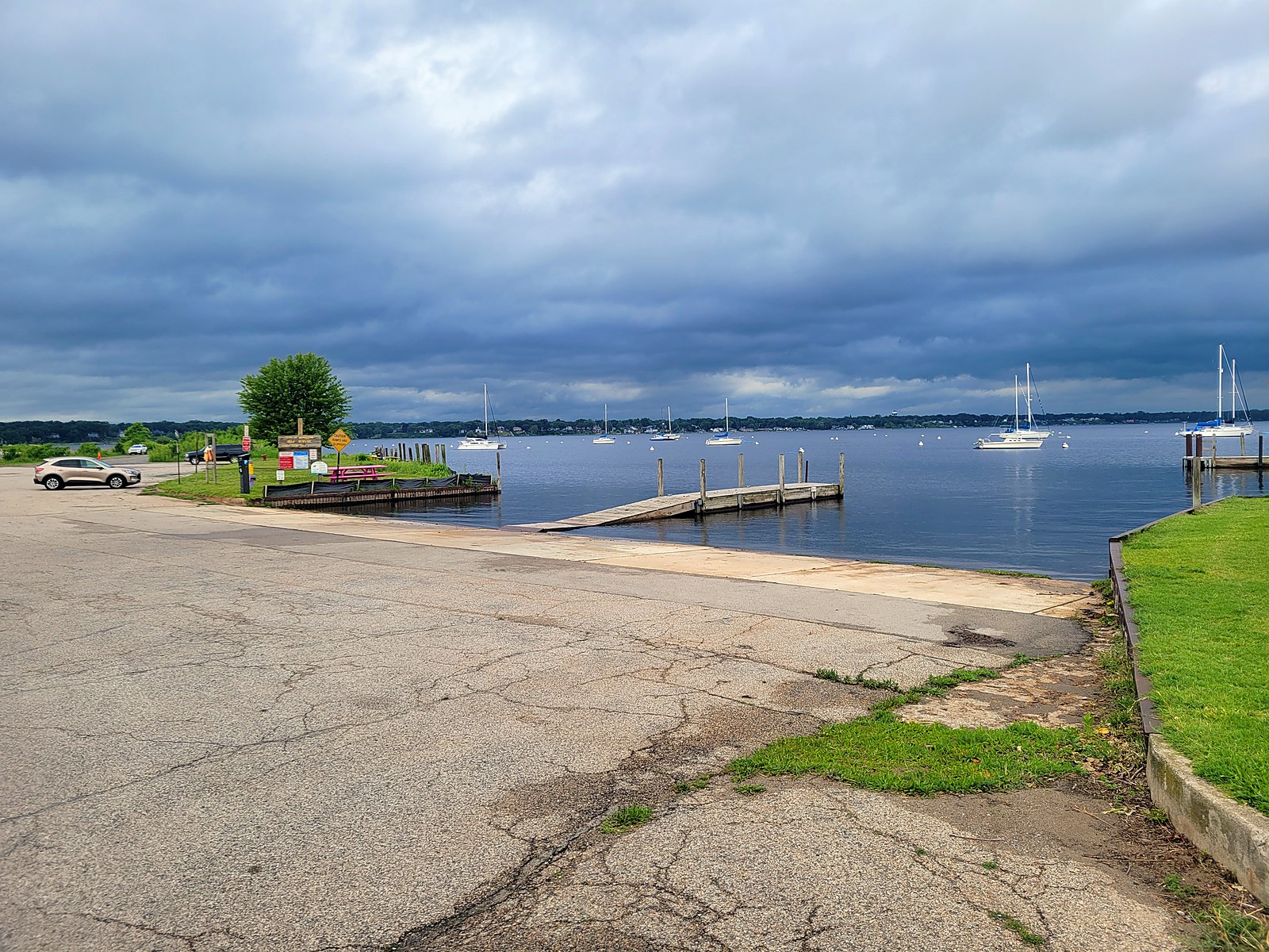 The Hartshorn launch ramp on Muskegon Lake, a wide concrete ramp leading to a courtesy dock, with sailboats moored on the open lake and the far shoreline beyond under an overcast sky.