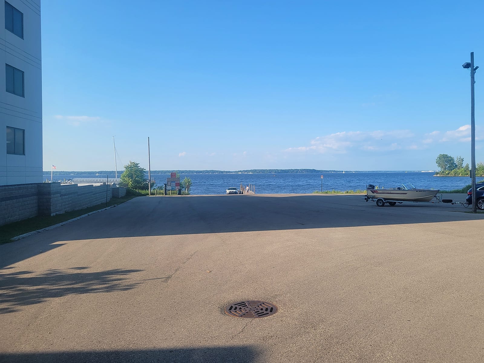The Jaycees (Cottage Grove) launch ramp on Muskegon Lake reopened, with the wide paved lot leading down to the ramp and courtesy dock, a vehicle at the water's edge, a fishing boat waiting on a trailer, and open water beyond on a clear summer day.