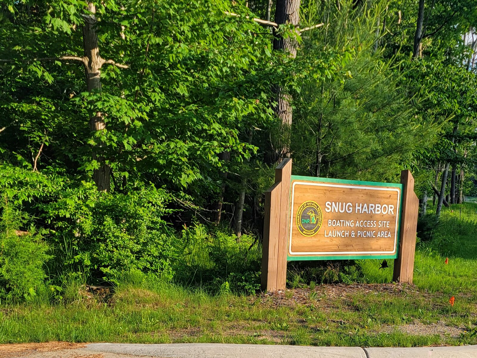 The Snug Harbor Boating Access Site entrance sign at the Muskegon State Park launch, reading Launch and Picnic Area, on a grassy median beside the park road.