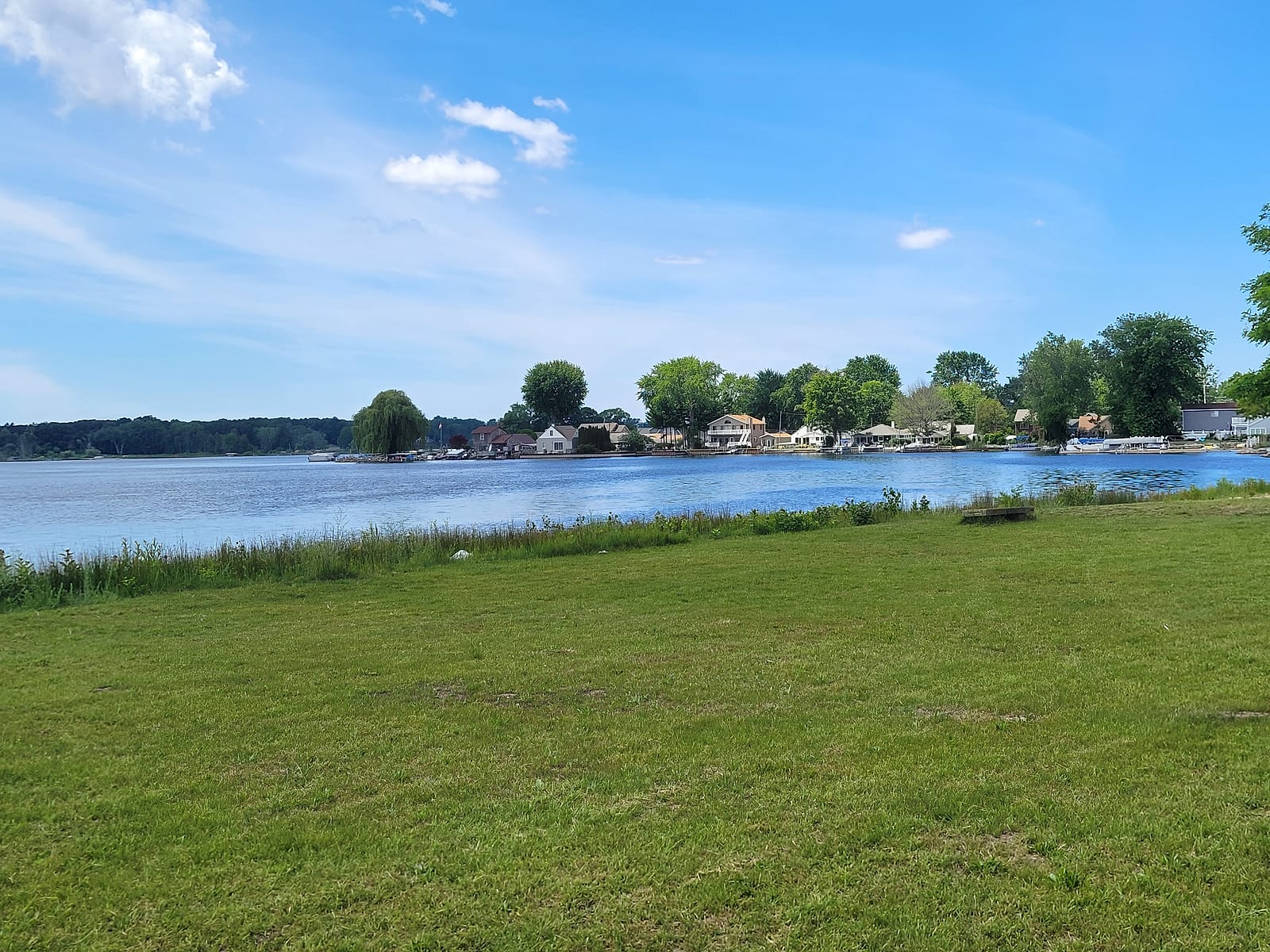 Mona Lake beside Airline Road in Norton Shores in June 2026, with waterfront homes, docks, and boats along the far shore under a summer sky