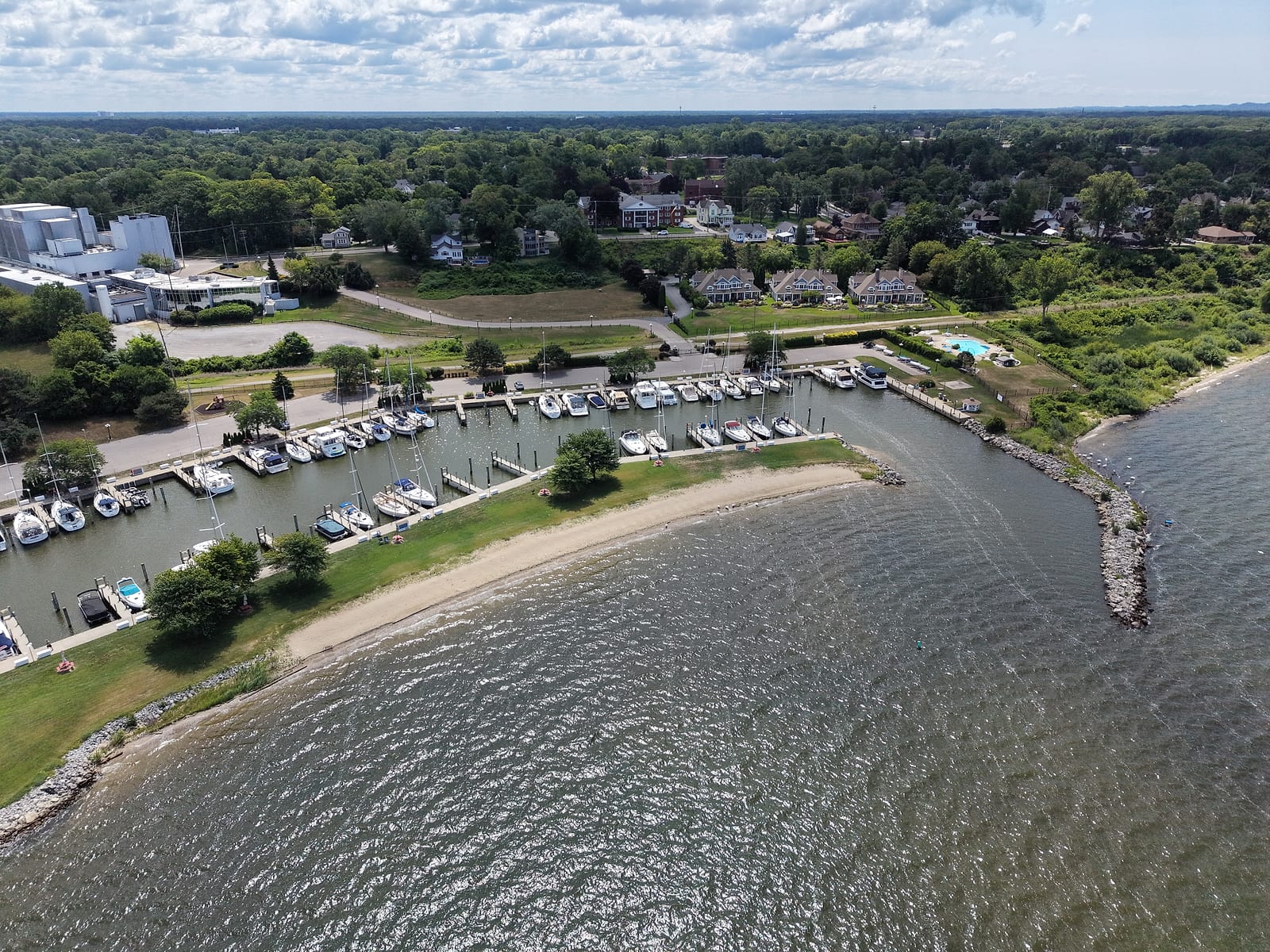 July 2026 aerial view of the rock-jettied harbor entrance and small beach at Lakeshore Yacht Harbour in Muskegon, with the Lakeshore Harbour townhomes and pool behind and the Nims neighborhood up the hill.