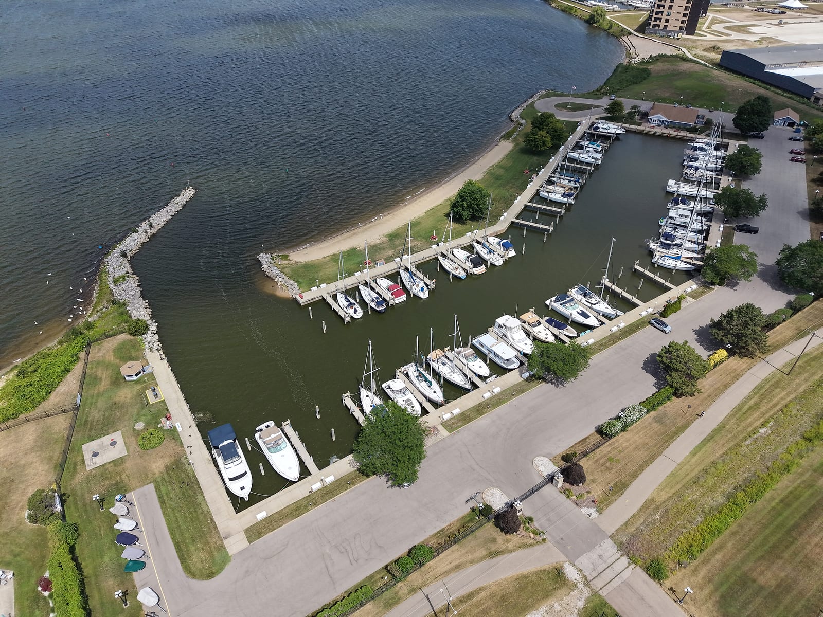July 2026 aerial view of Lakeshore Yacht Harbour in Muskegon: rows of owned boat slips inside a protected basin, the gated entrance drive, and the Muskegon Lakeshore Trail passing outside the fence.