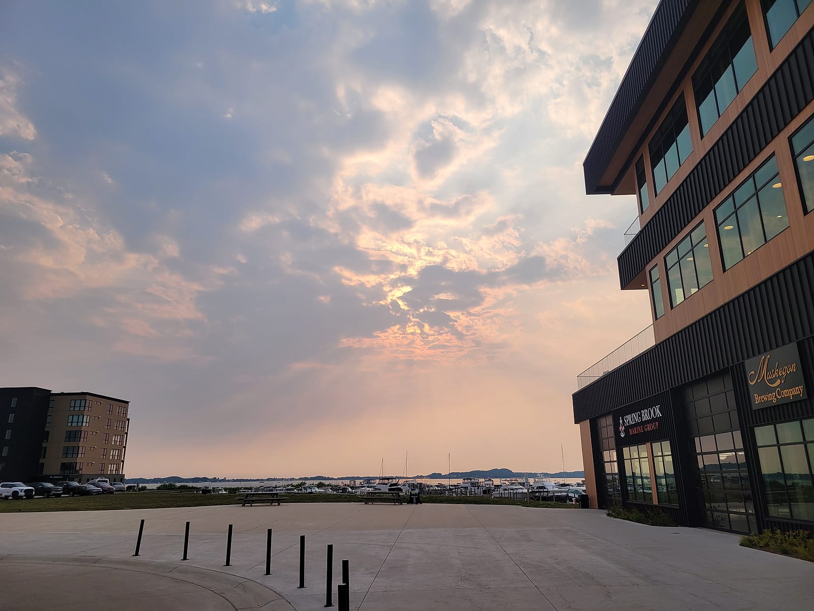 The mixed-use building at Adelaide Pointe in Muskegon at sunset in August 2026, with the Muskegon Brewing Company sign on its second floor, the condo building at left, and boats in the marina on Muskegon Lake