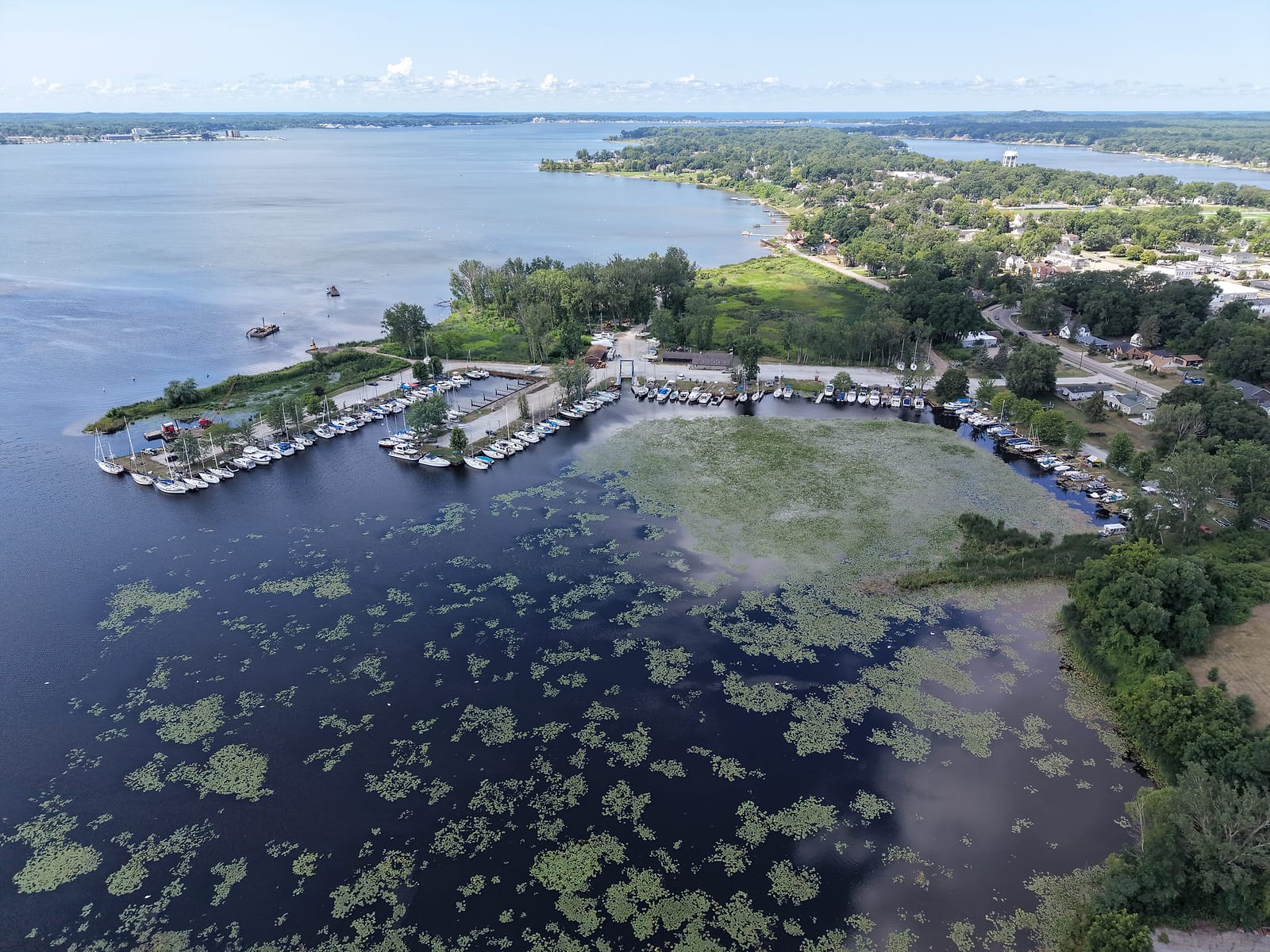 July 2026 aerial drone view of the Muskegon Conservation Club marina: members' boats in slips where the Muskegon River delta's lily-pad water opens onto Muskegon Lake, with a crane barge dredging the channel at the upper left.