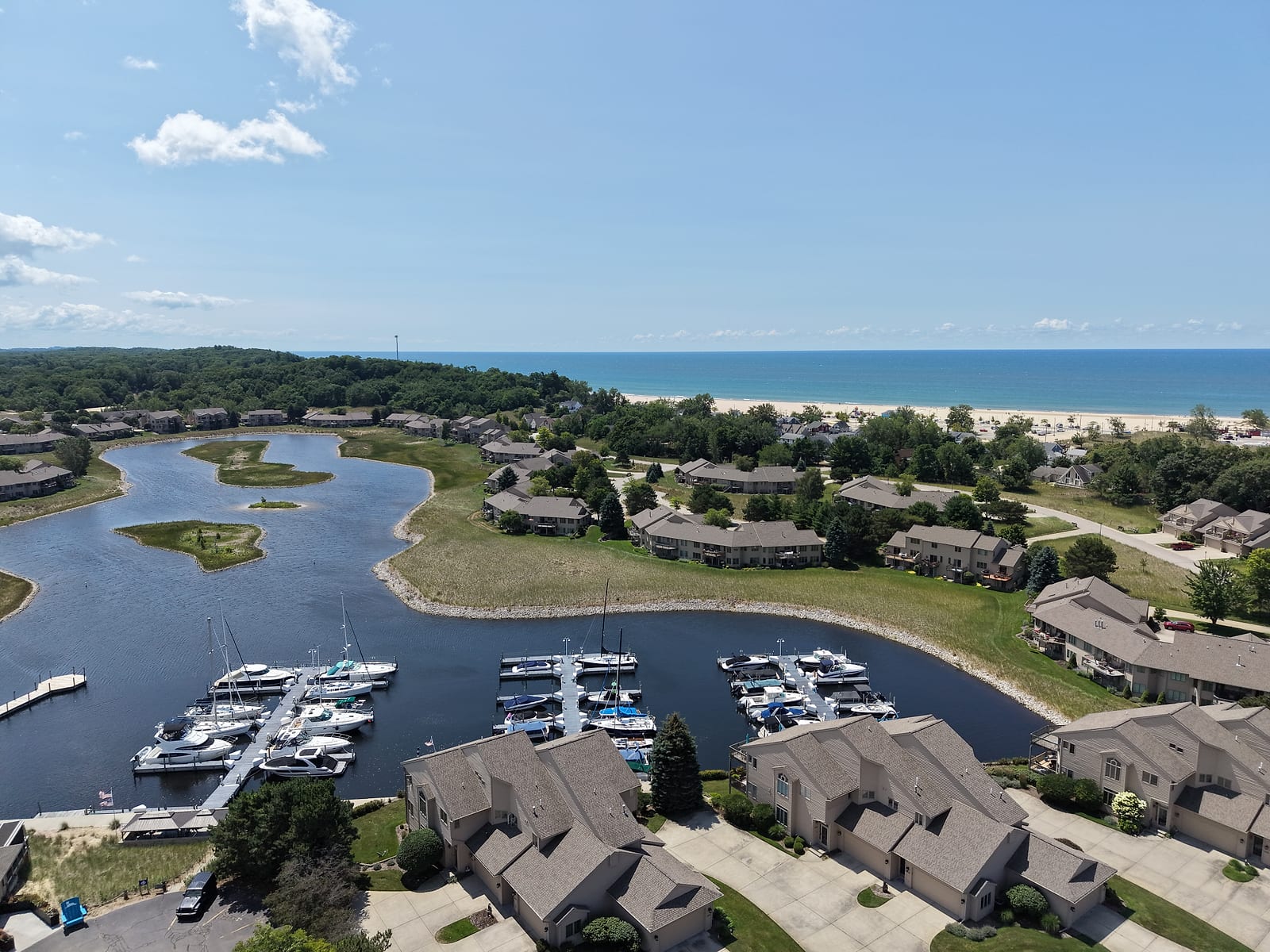 July 2026 aerial drone view of Pigeon Key Marina inside the Harbour Towne community in Muskegon: three floating docks of boat slips on a lagoon ringed by condo buildings, with Lake Michigan and its beach in the distance.