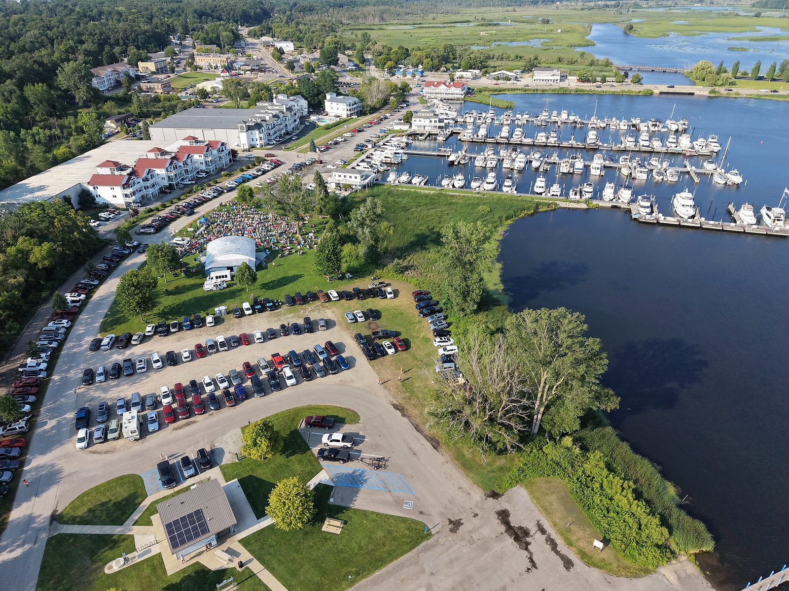 Aerial view of a summer concert crowd on the lawn at the Montague band shell, with full parking lots and the White Lake municipal marina behind