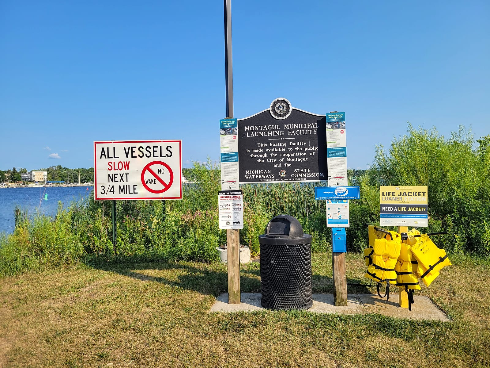 Sign cluster at the Montague boat launch: the Montague Municipal Launching Facility plaque, an all-vessels-slow no-wake sign, a muskellunge size-limit notice, and a stocked life-jacket loaner station